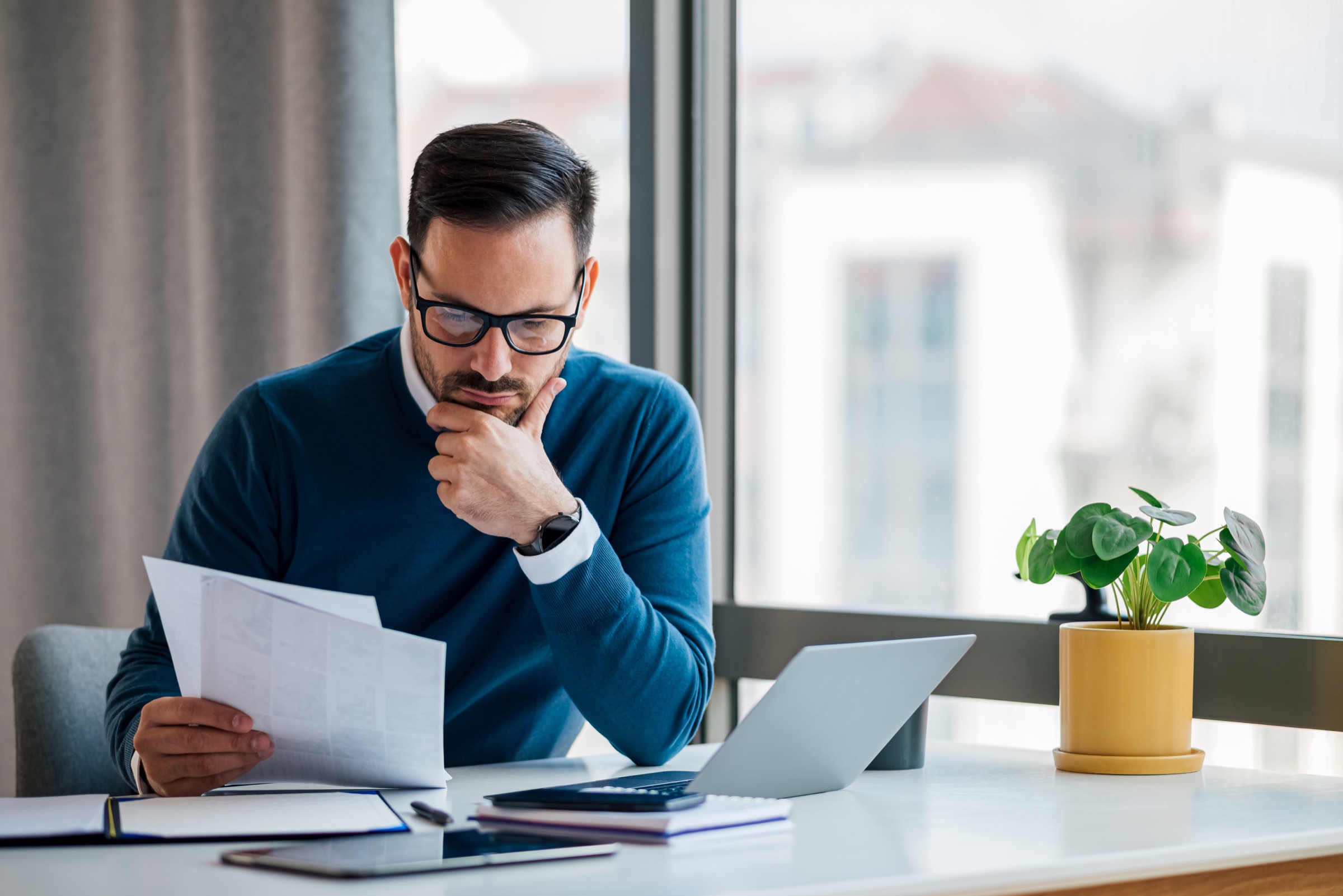 Man in glasses reviewing paperwork beside a laptop at a desk.
