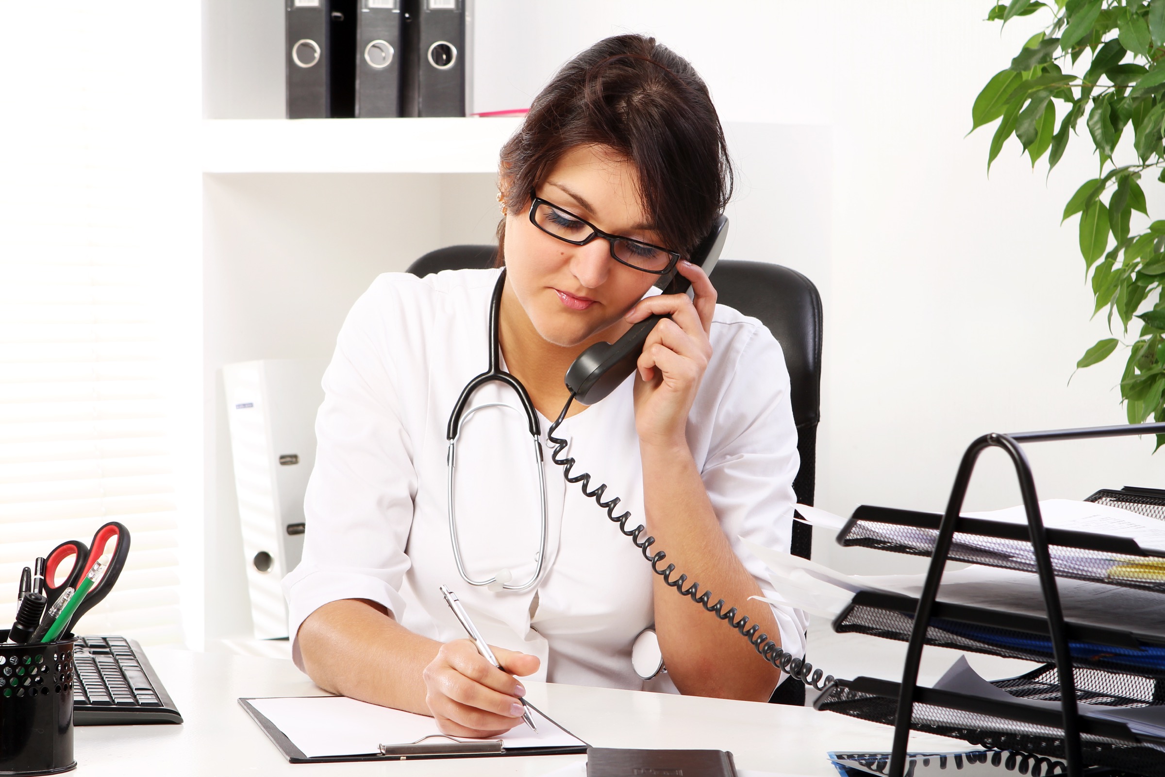 Healthcare professional writing on a clipboard while speaking on the phone.