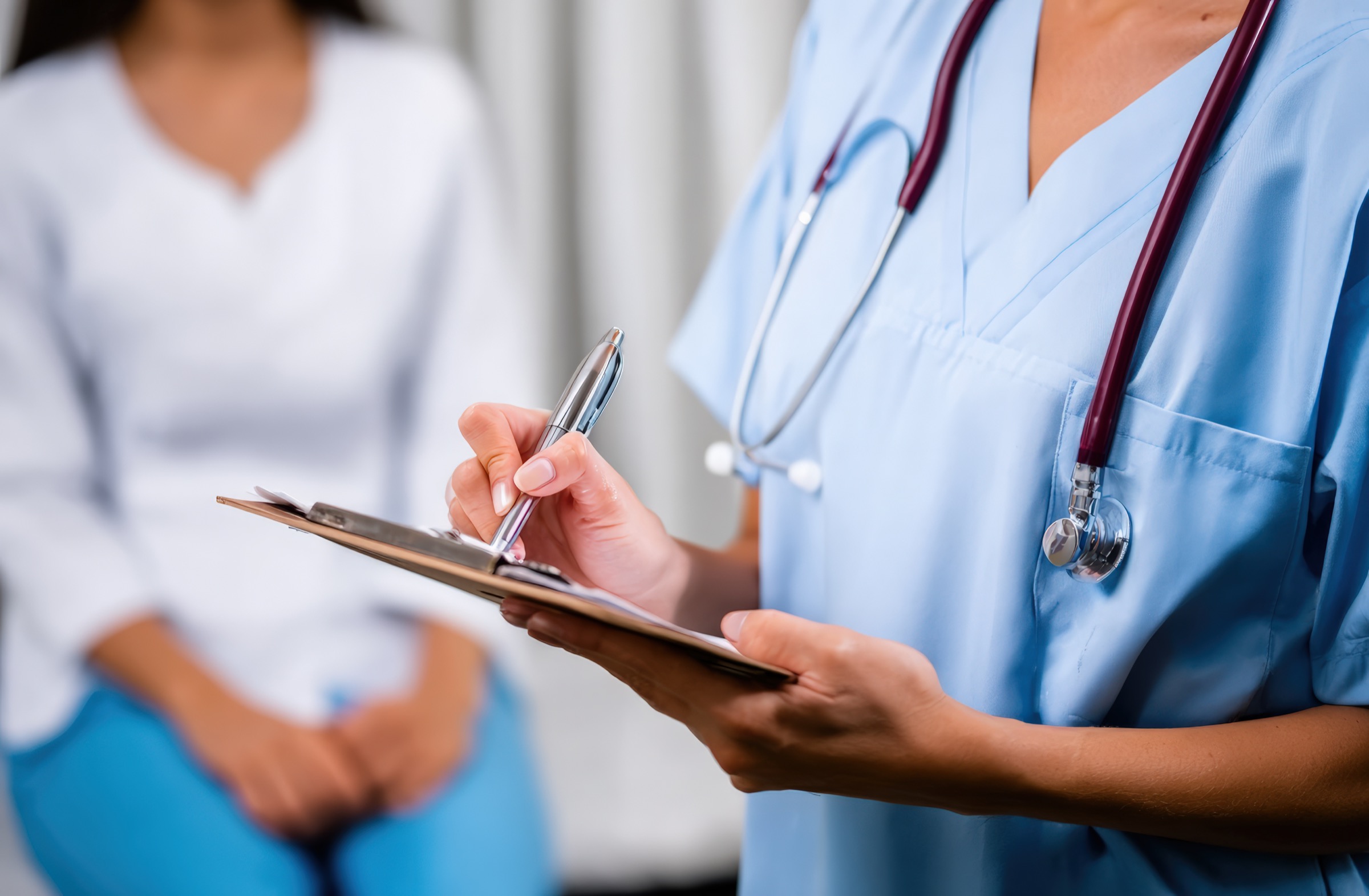 Healthcare professional in blue scrubs writing on a clipboard beside a seated person.