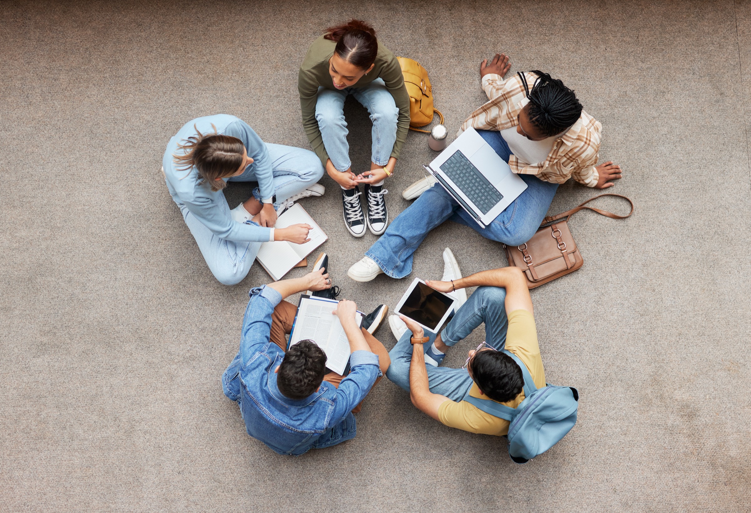 Overhead view of five people seated in a circle with a laptop, tablet, and notebooks.